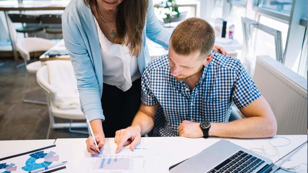 Two colleagues reviewing printed charts at a desk with a laptop, in a brightly lit office setting.