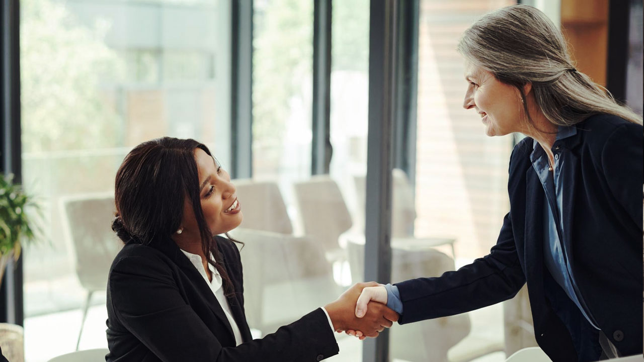 Two women in business attire shake hands in an office with large windows. One is seated, and the other stands leaning forward.